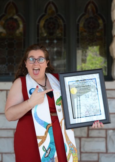 Pastor Mallory, a white woman with brown hair and glasses wearing a red dress and a white stole, holds a framed ordination certificate in one hand and points at it with the other hand. Her mouth is open in an exaggerated expression of excitement.